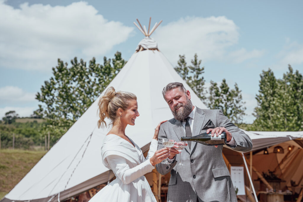 A joyful moment as the bride and groom pour champagne into glasses in front of their wedding tipi venue, set against a backdrop of rolling hills. The bride looks stunning in her long sleeve off-the-shoulder white gown, while the groom is dapper in his grey suit. The scenic beauty of the rolling hills adds a picturesque charm to the celebration.