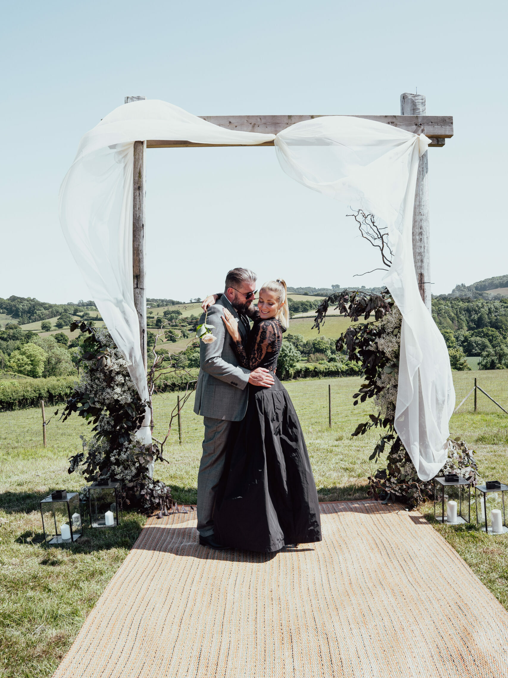 A bride and groom stand before a stunning wedding arch adorned with white drapes and modern black and white decor and flowers. The groom looks smart in a grey suit, while the bride is a vision of elegance in her long-sleeved black lace wedding dress. The backdrop features the rolling hills of Devon.