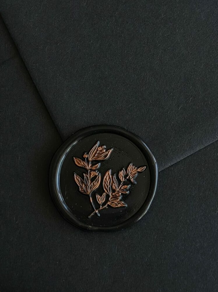 A handcrafted black botanical wax seal with delicate gold brushed detailing is elegantly affixed to a black envelope, showcasing intricate foliage detailing. The combination of the dark envelope and the exquisite botanical wax seal creates a captivating sense of sophistication and organic beauty, adding a touch of luxury to the wedding stationery.