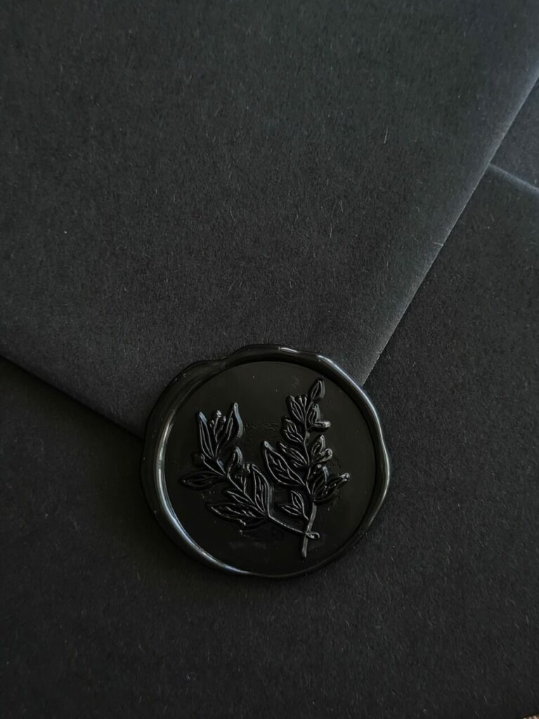 A handmade black botanical wax seal is elegantly affixed to a black envelope, featuring intricate foliage detailing. The combination of the dark envelope and the botanical wax seal creates a sense of sophistication and organic beauty.