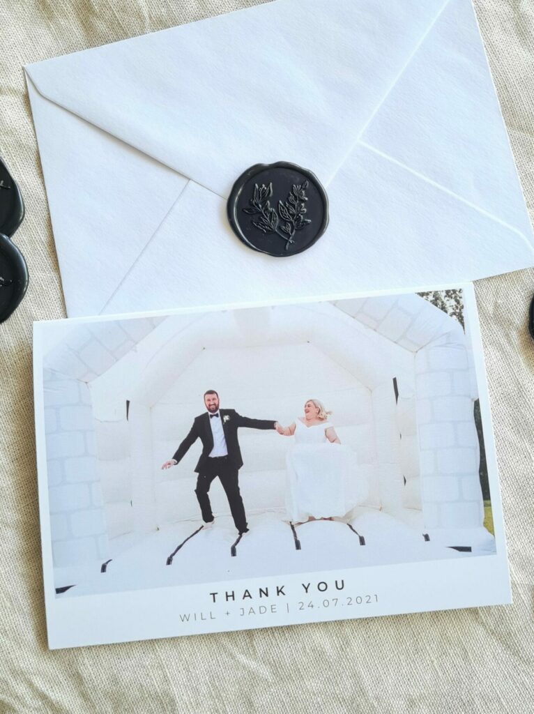 A wedding thank you card features a photo of the bride and groom with the word "Thank You" in elegant fonts, along with the couple's names and wedding date. The card is positioned next to a white envelope adorned with black wax seals, adding a touch of sophistication.
