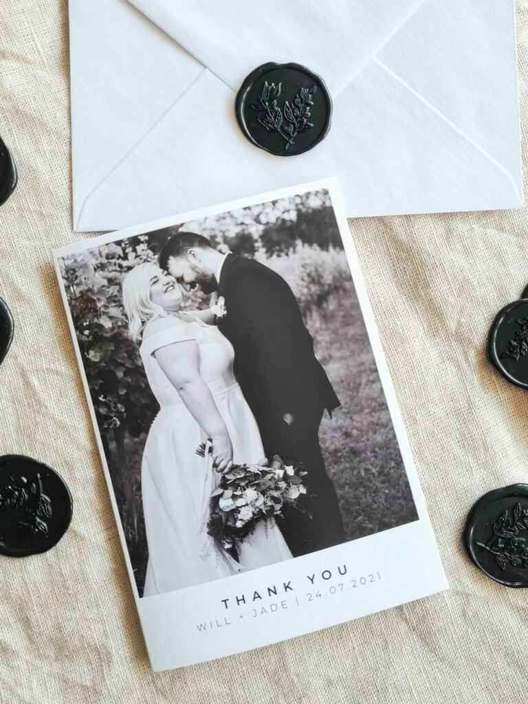 A wedding thank you card features a black and white photo of the bride and groom with the word "Thank You" in elegant fonts, along with the couple's names and wedding date. The card is positioned next to a white envelope adorned with black wax seals, adding a touch of sophistication.