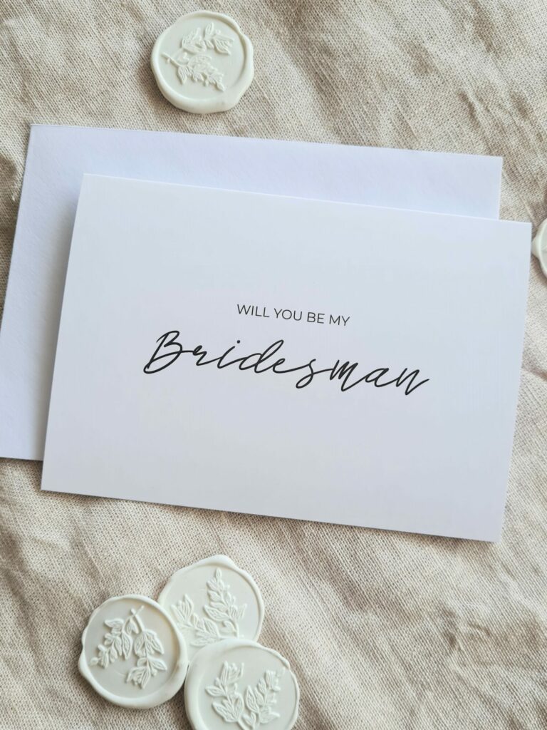 A white bridal party proposal card with the text "Will you be my Bridesman?" is elegantly arranged in modern minimal fonts. The card is placed on a beige linen background and is surrounded by white botanical wax seals, creating an aesthetic that is both contemporary and elegant.