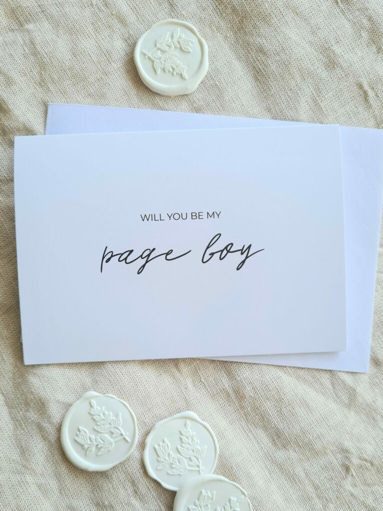 A white bridal party proposal card with the text "Will you be my Page Boy?" is elegantly arranged in modern minimal fonts. The card is placed on a beige linen background and is surrounded by white botanical wax seals, creating an aesthetic that is both contemporary and elegant.