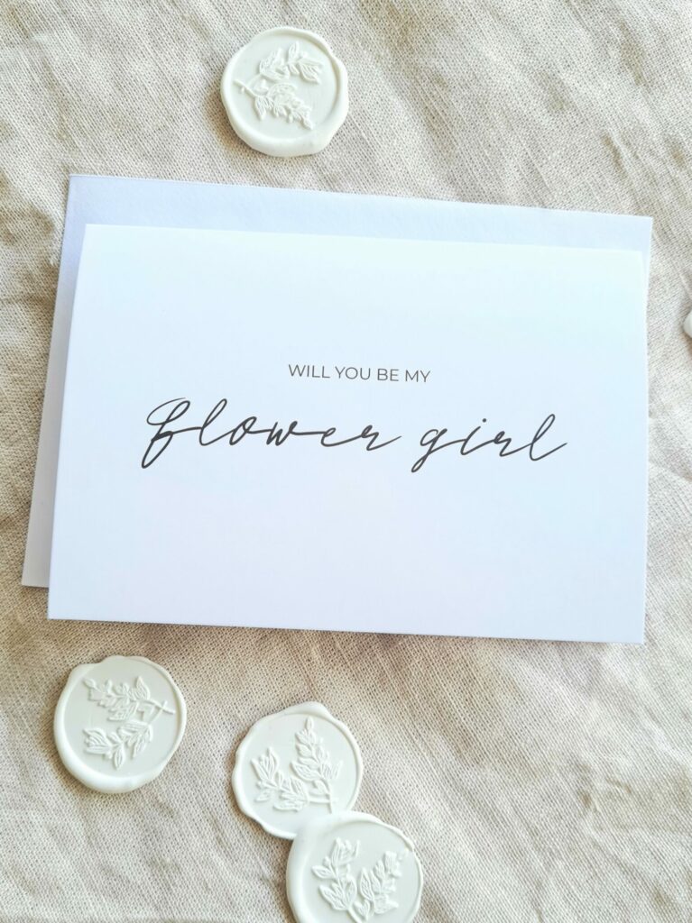 A white bridal party proposal card with the text "Will you be my Flower Girl?" is elegantly arranged in modern minimal fonts. The card is placed on a beige linen background and is surrounded by white botanical wax seals, creating an aesthetic that is both contemporary and elegant.