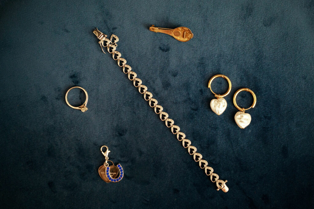 A collection of the bride's gold jewelry and trinkets captured on her wedding morning. The arrangement includes rings, earrings, a bracelet, and a brooch with a delicate horseshoe charm. These exquisite pieces are beautifully displayed on a rich blue velvet background, showcasing their elegance and significance for the special day.