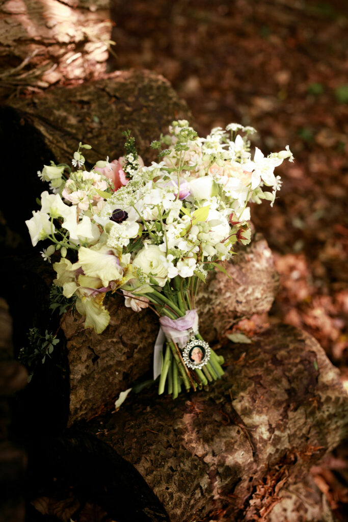 A stunning bridal bouquet in white, green, and pastel colors, delicately tied with a purple ribbon. A photo charm is placed within the bouquet. The bouquet is elegantly displayed against a rustic red brick wall, creating a beautiful contrast of colours and textures.