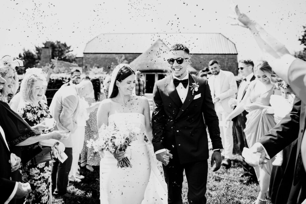 A timeless black and white image captures the newlywed couple walking down the wedding aisle after their ceremony. Their loved ones joyfully shower them with confetti. In the background, the picturesque Brickhouse Vineyard barn and roundhouse create a charming setting for this celebratory moment.