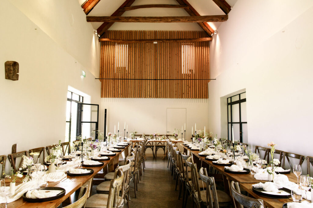 A modern and elegant table setting inside the barn at Brickhouse Vineyard near Exeter. The decor features a monochrome and neutral colour scheme, including flowers, creating a sophisticated and stylish ambiance. The table is beautifully set and prepared for the wedding breakfast, reflecting the attention to detail for this special occasion.