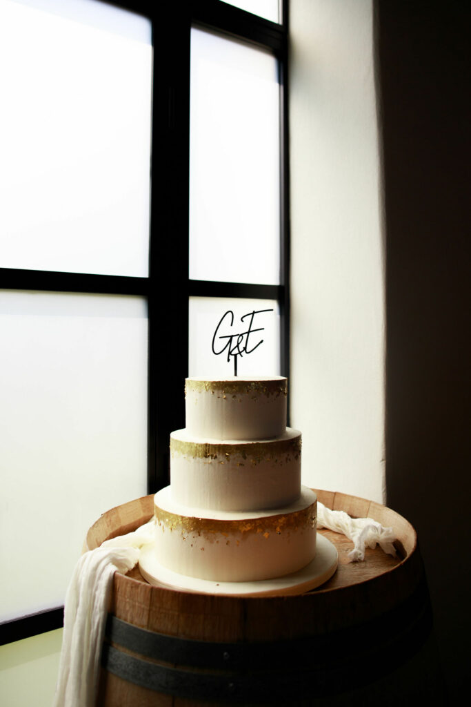 A modern and minimal wedding cake with three tiers, elegantly designed in white with delicate gold leaf detailing. The cake is topped with a cake topper that reads "G and E." It is displayed on a wine barrel and surrounded by white linen drapes, creating a stylish and sophisticated setup in front of the white and black windows of the barn at Brickhouse Vineyard.