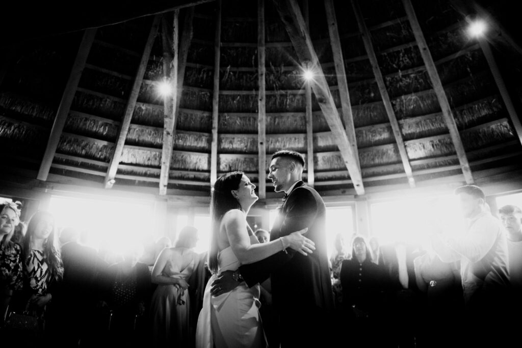 A black and white photo captures a couple sharing their first dance underneath the thatched roof of the roundhouse at Brickhouse Vineyard near Exeter. They are surrounded by their friends and family, creating a heart warming and intimate moment.