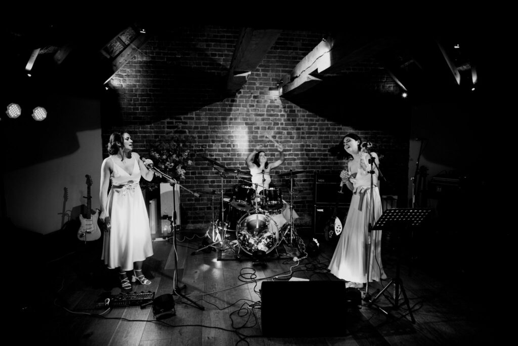 A black and white photo captures the bride playing the drums while surrounded by her bridesmaids who are singing inside the roundhouse at Brickhouse Vineyard. This musical and joyful moment reflects the celebration and camaraderie of the bridal party.