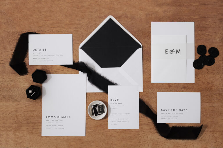 An assortment of wedding stationery items is neatly arranged on a wooden surface. The collection includes an RSVP card, a details card, and a save-the-date notice, all featuring elegant black and white design elements. A large envelope with a black interior sits prominently in the centre, accompanied by smaller envelopes and decorative black seals. The names ‘EMMA & MATT’ are visible on several pieces, indicating the couple for whom this stationery set is intended. Black fabric ribbon pieces add texture to the composition.