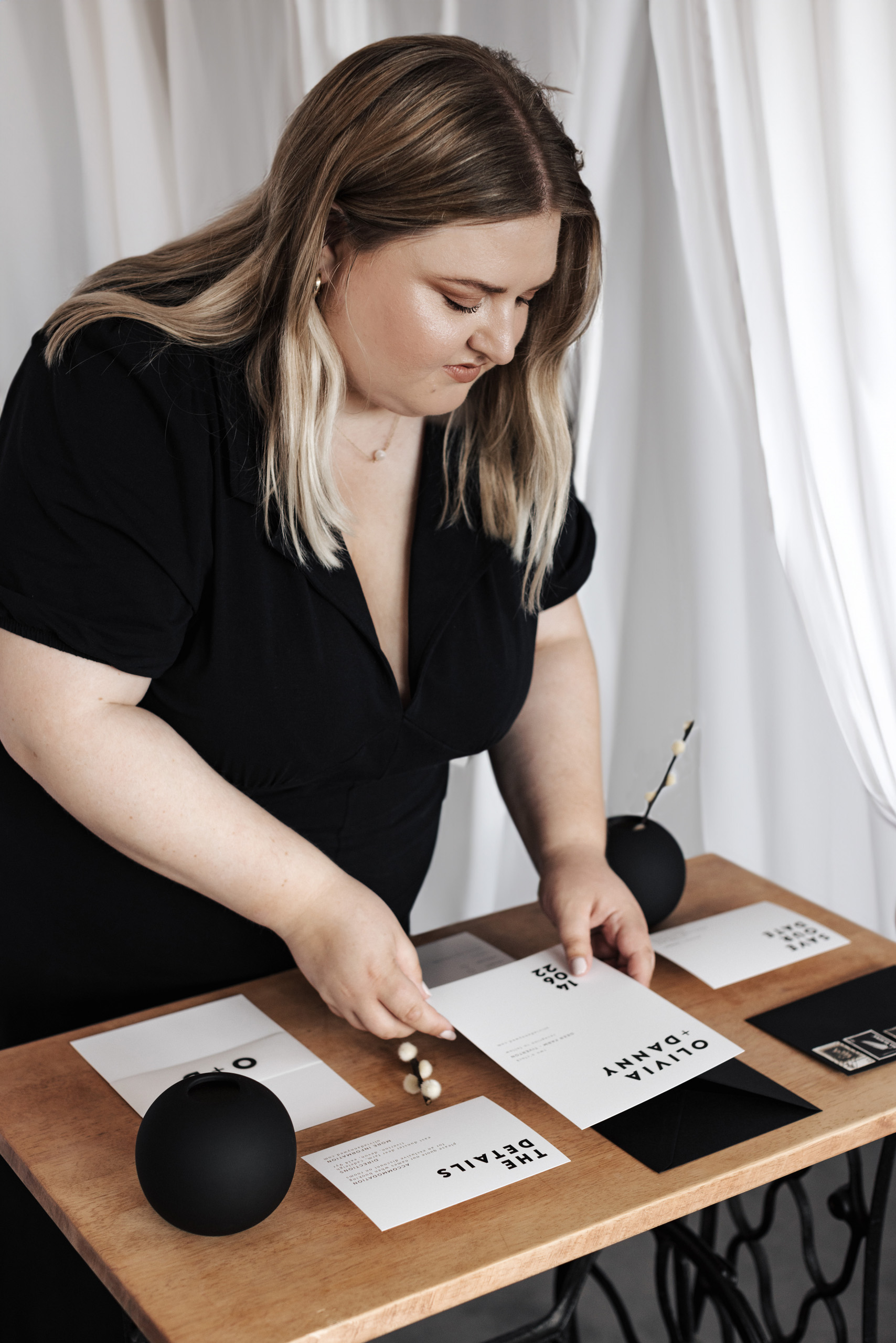 A person is stood over a table with their hands arranging various wedding stationery cards on a wooden surface. The cards feature different black text on a white background. There is an invitation, save the date card, rsvp card and a belly band which are surrounded by black and white decorative items such as envelopes, vases and dried stems.