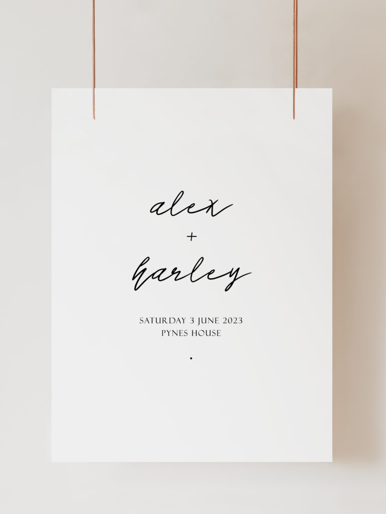 An image of a black and white welcome sign featuring modern calligraphy-style text that reads "alex + harley" with the date and venue below. The sign is hanging on ribbon with a beige background behind.