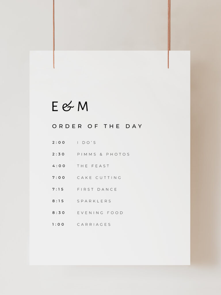 A white wedding order of the day sign with modern black text reading "E and M Order of the Day" with the timings and events listed below. The sign is hanging on ribbon with a beige background behind.