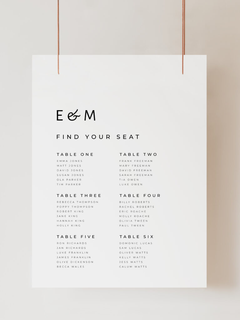 A white wedding table plan sign with modern black text reading "E and M Find Your Seat" with 6 table numbers with guests listed below. The sign is hanging on ribbon with a beige background behind.