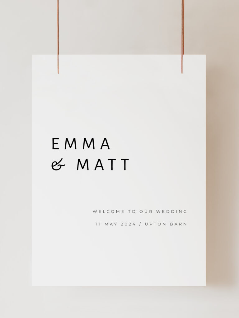 A white wedding welcome sign with modern black text reading "Emma and Matt Welcome to our Wedding" with the wedding date and venue below. The sign is hanging on ribbon with a beige background behind.