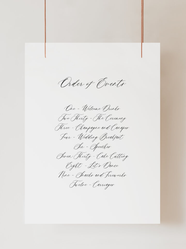 A white Order of the Day Sign with cursive handwriting. The text reads ‘Order of Events' with timings and a list of events for the day. The elegant script and structured list suggest it is a wedding program outlining the sequence of events for the day. The sign is hanging on ribbon with a beige background behind.