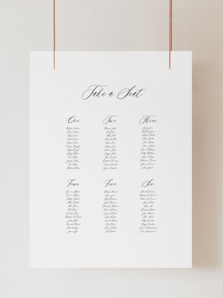 A white seating plan sign with the title ‘Take a Seat’ at the top in cursive font. Below are six columns of names, each column headed by a table number from one to six, also written in cursive. The names are listed in a smaller font, arranged neatly under each table number indicating seating arrangements. The sign is hanging on ribbon with a beige background behind.