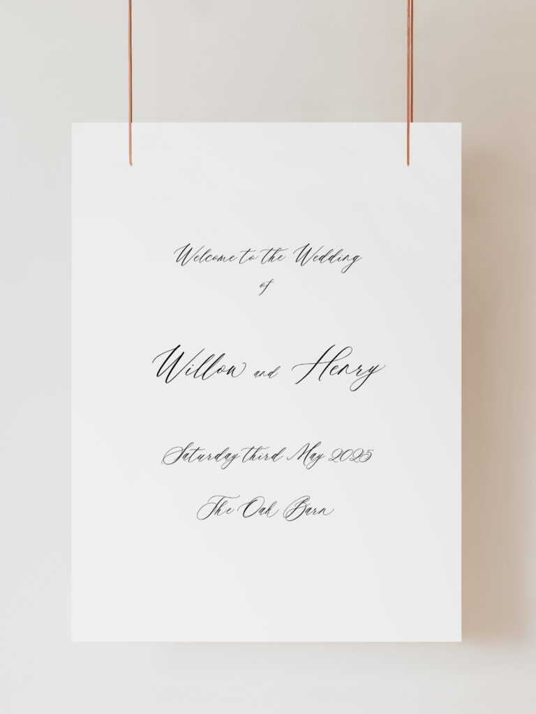 A white wedding welcome sign with cursive handwriting is suspended against a plain wall by two copper-colored strings at the top corners. The handwritten text reads ‘Welcome to the Wedding of Willow & Henry, Saturday Third May 2025, The Oak Barn’ The sign is hanging on ribbon with a beige background behind.