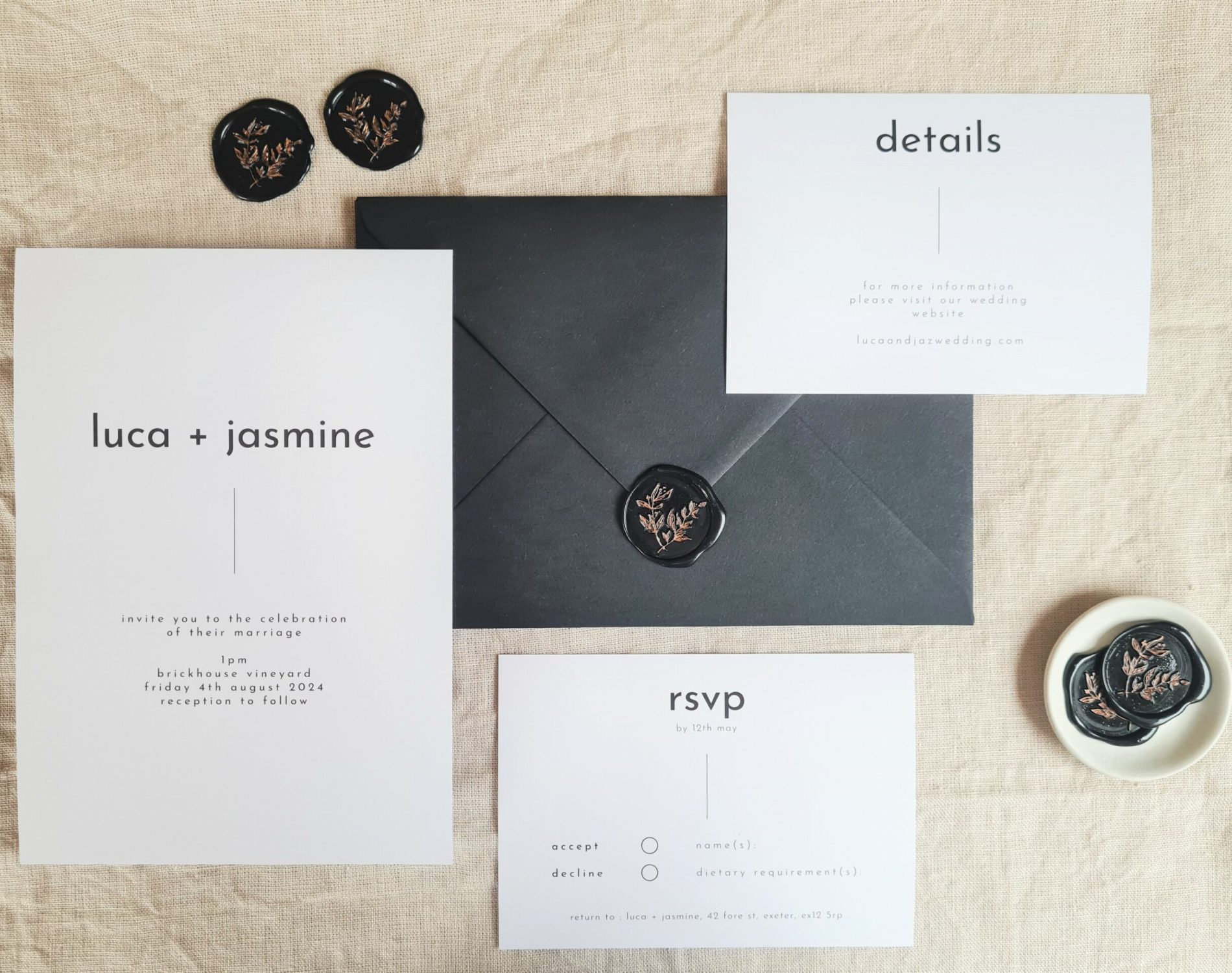 A black and white invitation suite flat lay. There is an RSVP card, invite and Details card. They are set against a beige linen background. Black wax seals, a mini white plate and a black envelope is arranged around it.