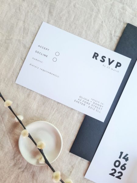 A black and white RSVP card is set against a beige linen background. Modern dried flowers are arranged around it and a white mini plate and a black envelope.