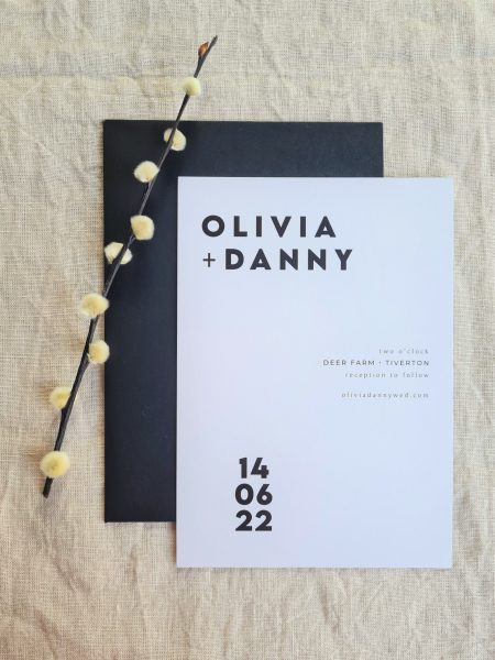 A black and white invitation with the words "Olivia and Danny" written on it. The card is set against a beige linen background. Modern dried flowers are arranged around it and a black envelope.