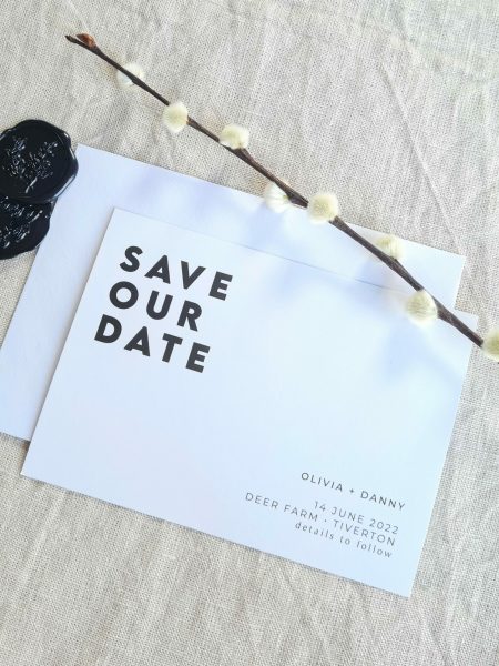 A black and white save the date card with the words "Save our Date" written on it. The card is set against a beige linen background. Modern dried flowers are arranged around it and black wax seals with botanical detailing.