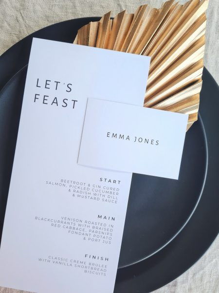 A black and white wedding menu with the text "Let's Feast" and a place card are placed on a black plate. The plate is set on a beige linen background and is accompanied by a gold leaf fan. The overall aesthetic is elegant and sophisticated.