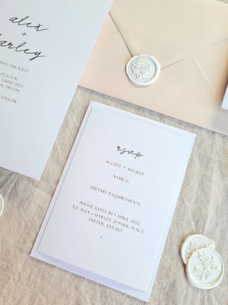 A black and white RSVP card is placed on a beige linen background and is accompanied by natural dried floral elements, white botanical wax seals and a beige envelope. The overall aesthetic is elegant, modern and sophisticated.