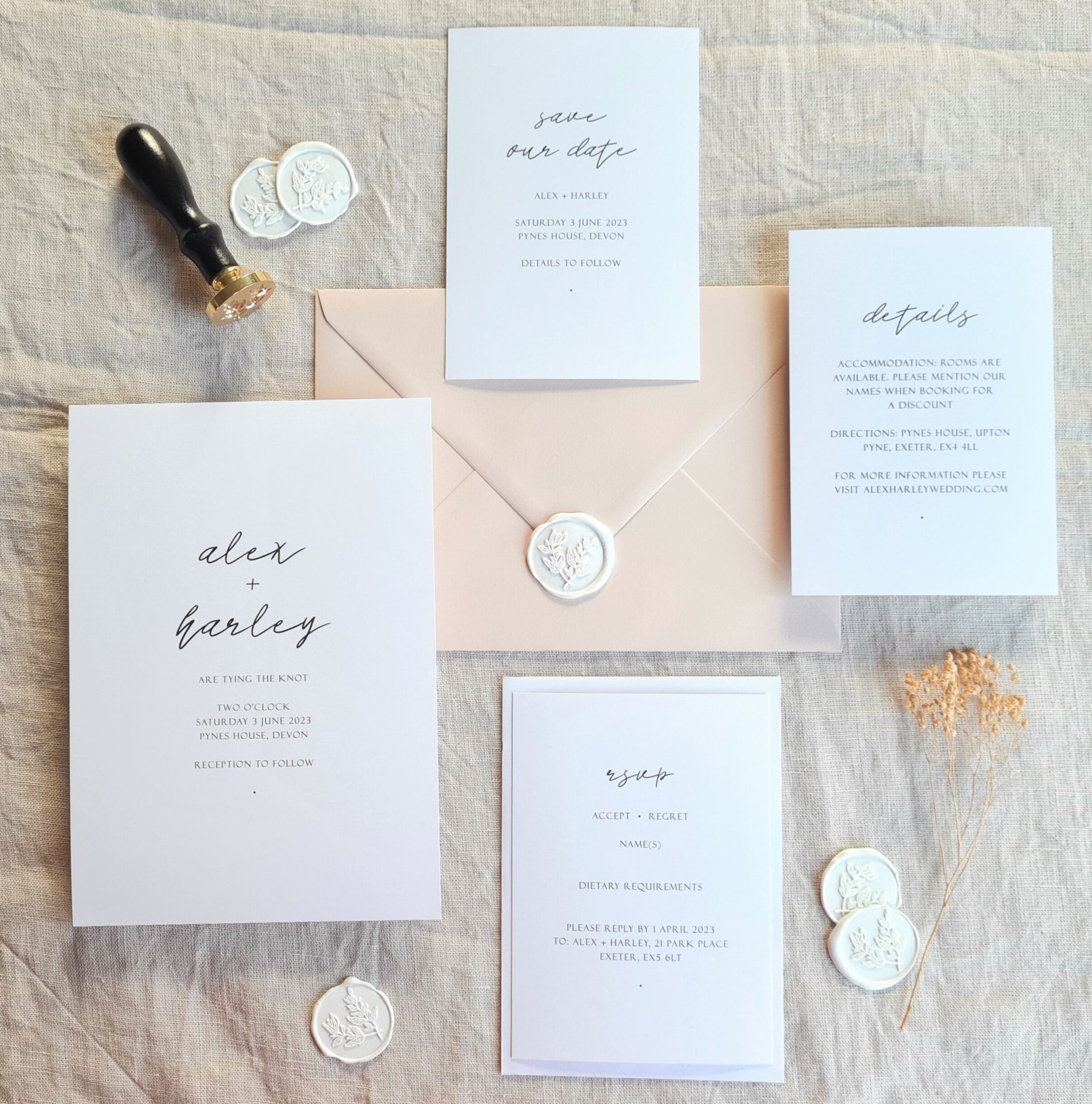 This is a flat lay image of a black and white modern wedding invitation suite set on a beige linen background with various dried flowers scattered around it. The suite includes a save the date card, an invitation, an RSVP card, and a details card. The save the date card is a simple black and white design, while the invitation features bold black text on a white background, with a black and gold wax stamp and white wax seals.