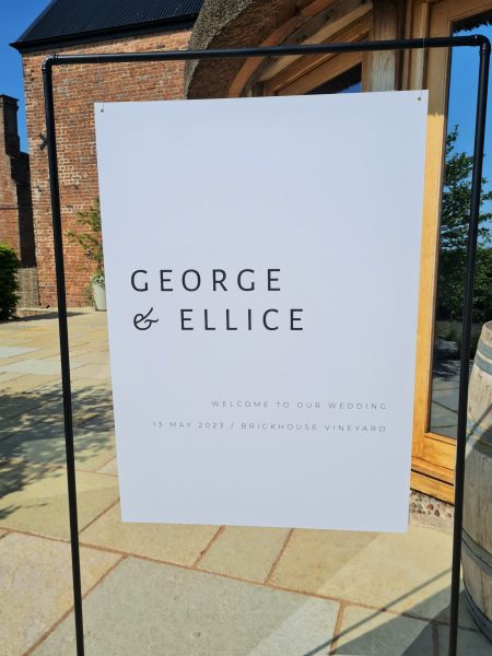 A black and white minimal and modern wedding welcome sign placed in front of the stunning red brick barn and thatched roundhouse at Brickhouse Vineyard in Exeter, Devon. The sign is made of matte black copper and features sleek and contemporary typography. The contrast between the black sign and the red brick barn creates a visually striking image.