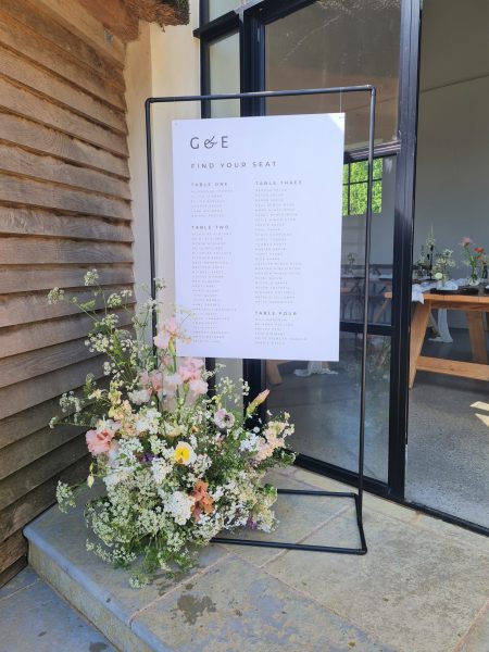 A black and white minimal and modern wedding welcome sign made of matte black copper placed in front of the breathtaking red brick barn and thatched roundhouse at Brickhouse Vineyard in Exeter, Devon. The sign features sleek and contemporary typography. In front of the sign, on the floor, there is a stunning modern floral arrangement. The arrangement showcases a mix of pink, green, white, and orange flowers, creating a vibrant and natural color palette. The combination of the sleek sign and the vibrant floral arrangement adds a touch of elegance and charm to the scene.