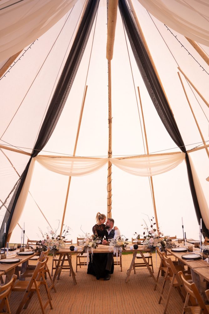 The bride and groom sit together at the head of the beautifully styled wedding table inside a beige wedding tipi. The table is elegantly dressed with modern monochrome and neutral styling, including stationery. The groom looks smart in his grey suit, while the bride is stunning in her long-sleeved black lace wedding dress. Their presence adds a touch of sophistication to the chic and contemporary table setting.