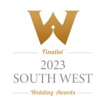 The image features a wedding badge for the “2023 South West Wedding Awards.” At the top, there is a large letter ‘W’ in a serif font, representing the word ‘Wedding.’ Below this, the word ‘Finalist’ is written in an elegant script. A horizontal line separates ‘Finalist’ from the year ‘2023,’ which is presented in bold, sans-serif numerals. Underneath, ‘SOUTH WEST’ appears in capital letters with a smaller font size than ‘2023,’ and below that, ‘Wedding Awards’ is written in a larger serif font. The colour scheme includes shades of gold and white, suggesting prestige and celebration associated with being an award finalist.