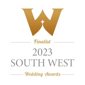 The image features a wedding badge for the “2023 South West Wedding Awards.” At the top, there is a large letter ‘W’ in a serif font, representing the word ‘Wedding.’ Below this, the word ‘Finalist’ is written in an elegant script. A horizontal line separates ‘Finalist’ from the year ‘2023,’ which is presented in bold, sans-serif numerals. Underneath, ‘SOUTH WEST’ appears in capital letters with a smaller font size than ‘2023,’ and below that, ‘Wedding Awards’ is written in a larger serif font. The colour scheme includes shades of gold and white, suggesting prestige and celebration associated with being an award finalist.