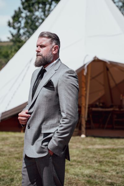 The groom stands in front of a white wedding tipi. He is impeccably dressed in a grey wedding suit with a black tie and other stylish details. His attire exudes sophistication and elegance against the backdrop of the white tipi, setting the scene for a memorable wedding celebration.