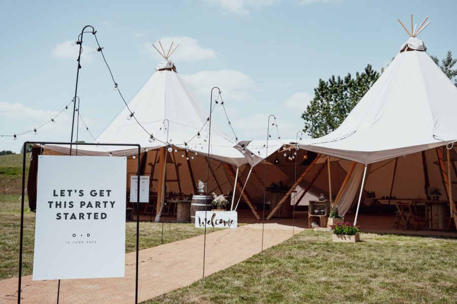 A black and white wedding welcome sign reading 'Let's get this party started' displayed in front of tipis at Deer Farm Tipi Weddings near Exeter. The sign is designed in a classic and elegant black and white colour scheme, with clear and bold text that catches the eye. The sign is displayed in front of the tipis, which are a unique and stylish venue choice for a wedding.