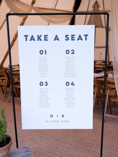 A black and white table plan hanging within a black copper sign frame that reads "Take a seat", inside a tipi. The sign features minimalist typography with black and white colours, providing a modern and stylish aesthetic.
