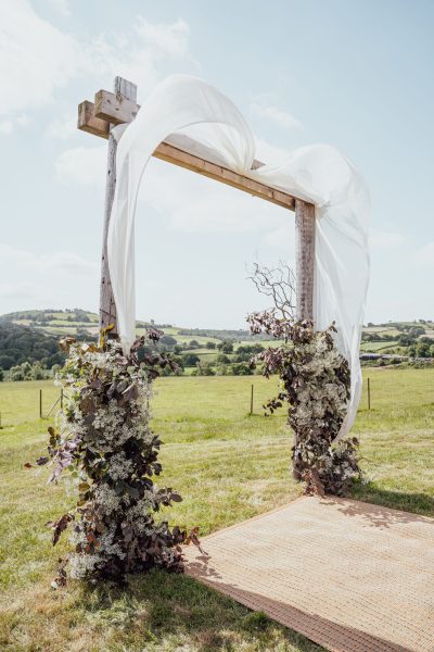 A wooden arch stands at the end of the wedding aisle, adorned with contemporary white drapes and neutral flowers. The arch serves as a beautiful focal point against the backdrop of the serene countryside setting, creating a picturesque scene for the wedding ceremony.