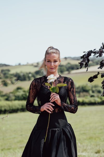 A bride stands gracefully in front of the picturesque Devon rolling hills. She is wearing a stunning black long-sleeved lace wedding dress and holds a single white rose. The contrast between the bride's attire and the natural beauty of the landscape creates a striking and memorable image.