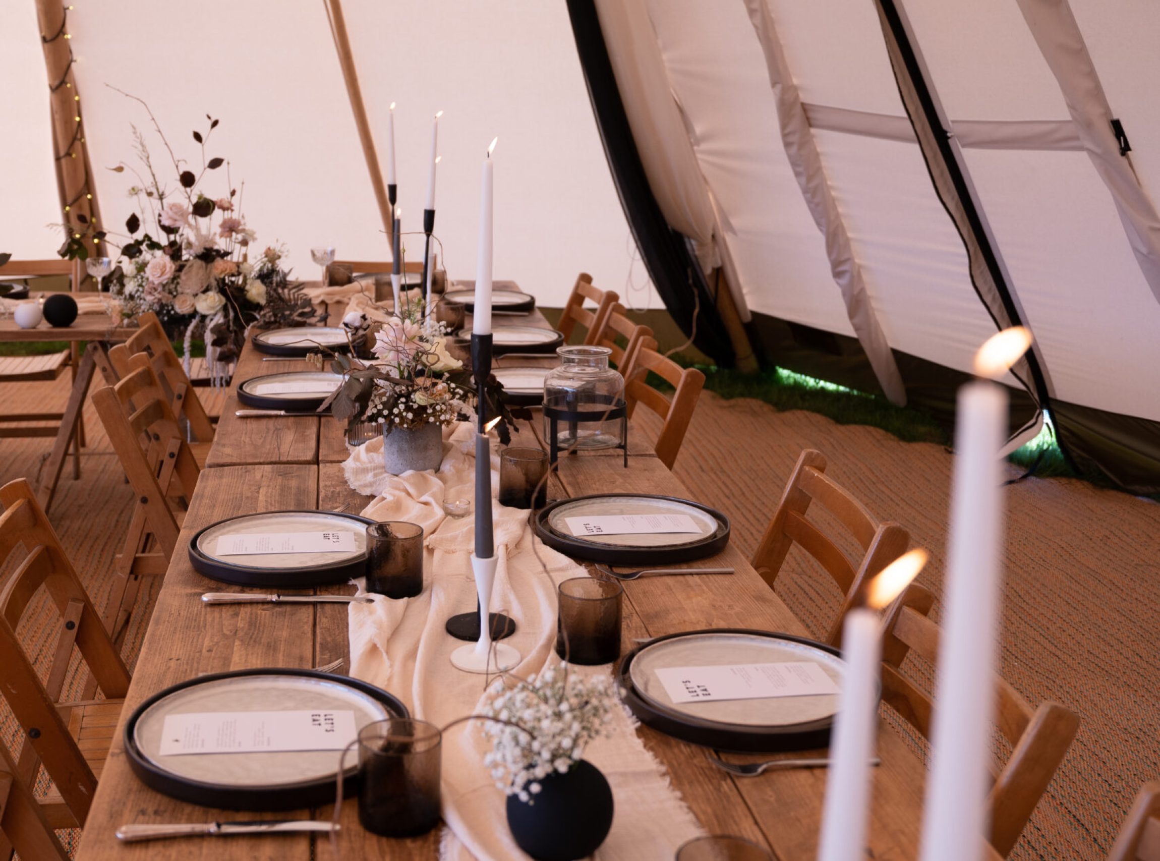 A black and white wedding menu tables cape with beige décor displayed on a table inside a tipi at Deer Farm Tipi Weddings near Exeter. The wooden table features a beige tablecloth, modern black and grey plates with a black and white floral accent and candles.