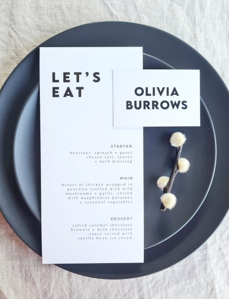 A black and white place card and menu with the words "Let's Eat" written on it. The card and menu are placed on a black plate and are set against a beige linen background. Modern dried flowers are arranged on the plate.