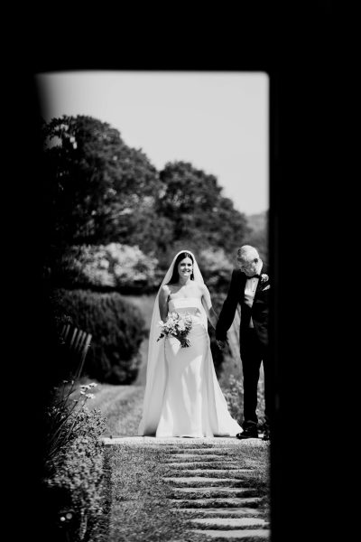 A timeless black and white image captures the moment as the bride is walked down the aisle by her father. The bride, dressed in a minimal and elegant white wedding dress and veil, holds a bouquet. This classic and emotional moment is beautifully depicted in monochrome tones.