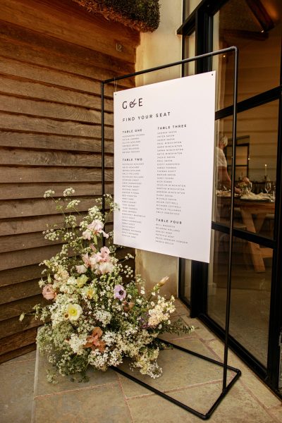 A modern and minimal wedding seating plan sign in monochrome, featuring classic typography, is elegantly displayed on a black copper sign frame. The sign is positioned in front of the barn at Brickhouse Vineyard, with a white and pastel floral arrangement adding a touch of elegance to the scene.