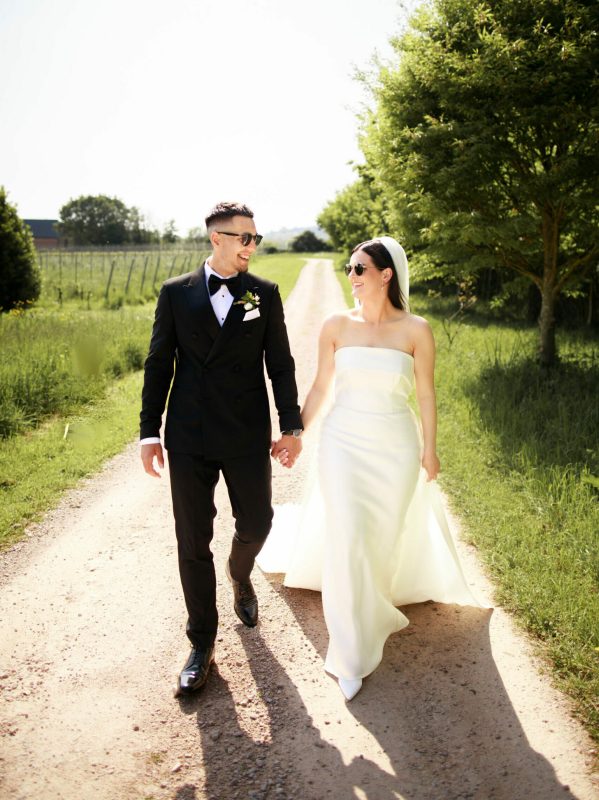 A beautiful moment captured as the bride and groom walk down the driveway of Brickhouse Vineyard, hand in hand. The bride is dressed in an elegant strapless white wedding gown, while the groom looks sharp in a black tuxedo. In the background, the scenic vineyard landscape adds to the romantic atmosphere of the moment.