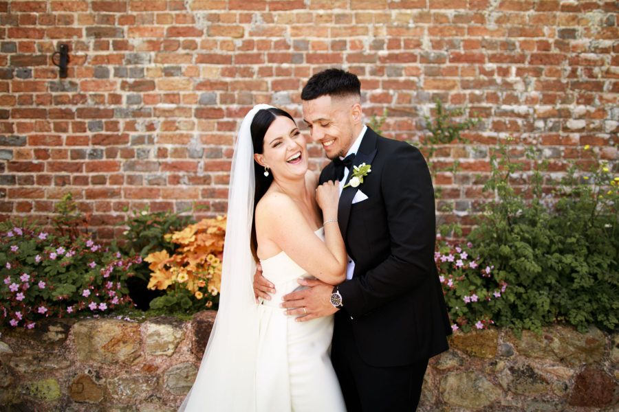 A delightful photo captures the bride and groom sharing laughter as they stand in front of the picturesque Brickhouse Vineyard barn. In the background, colourful flower beds add to the natural beauty of the scene, creating a joyful and charming moment.