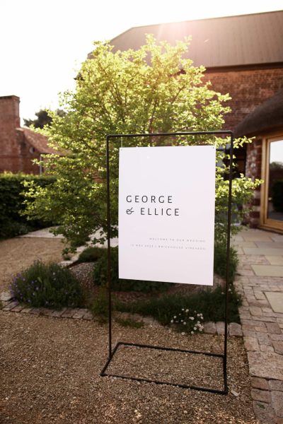 An elegant and modern minimal wedding welcome sign in monochrome with classic contemporary typography. The sign is elegantly displayed within a black copper sign frame. It stands in front of the barn and roundhouse at Brickhouse Vineyard near Exeter in Devon, creating a stylish and welcoming entrance for the wedding celebration.