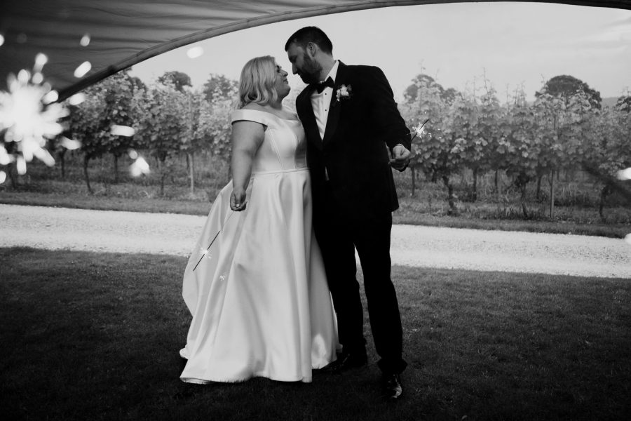 A black and white image of a bride wearing a white wedding dress and standing next to the groom in a black tie suit. They are standing in the vineyards of Brickhouse Vineyard near Exeter, Devon, with grape vines visible in the background. Both the bride and groom are holding sparklers.